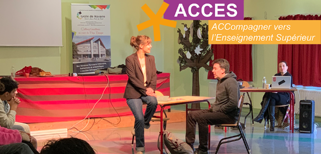 Une femme assise sur une table parle avec un homme assis sur une chaise. En arrière plan une femme assise devant un ordinateur